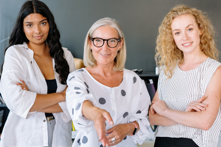 Three women standing and smiling, with one in the center extending her hand for a handshake. 
