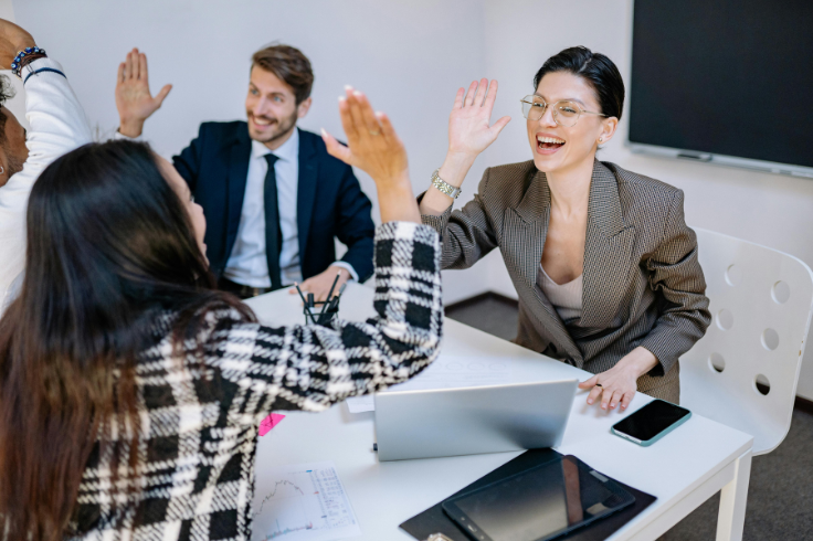 Four colleagues in business attire give each other a high five around a table in a modern office setting. 