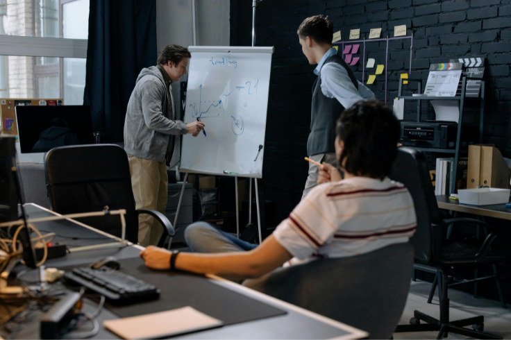 A person draws a graph on a whiteboard while two others watch in an office setting.