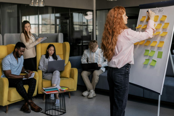 A team is gathered in a modern office space, with one person writing on a whiteboard covered in sticky notes while others work on laptops or take notes. 