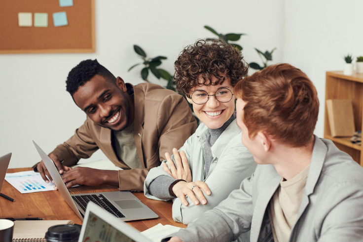 Three colleagues are smiling and looking at each other while working on laptops at a wooden table. Two laptops are visible, along with charts and papers on the table.
