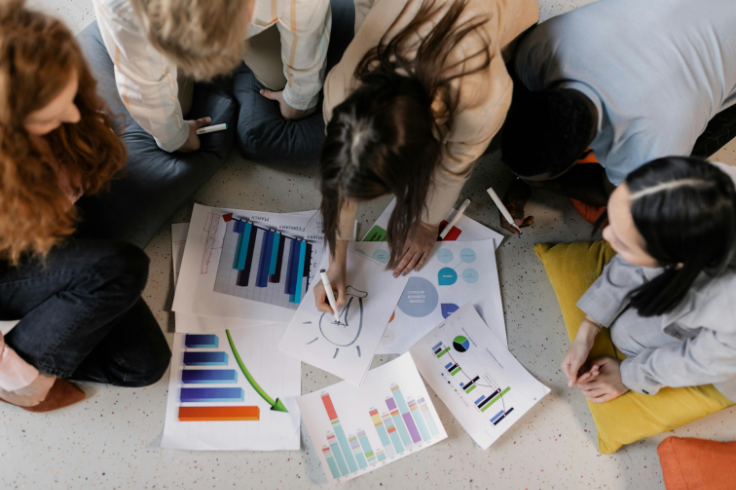A group of people are gathered around charts and graphs spread on the floor, engaged in a collaborative discussion. 