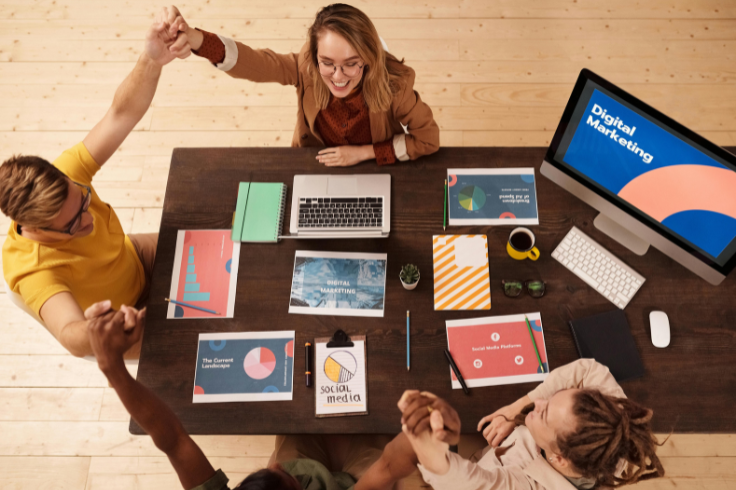 A team high-fiving over a table filled with marketing charts, graphs, and a laptop, with a computer monitor displaying 'Digital Marketing'.