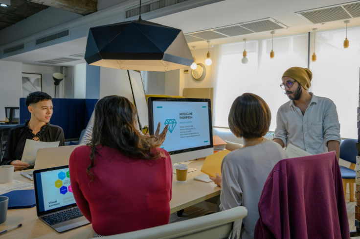 A group of people in an office collaborating around a desk with laptops and a computer displaying presentation slides. 