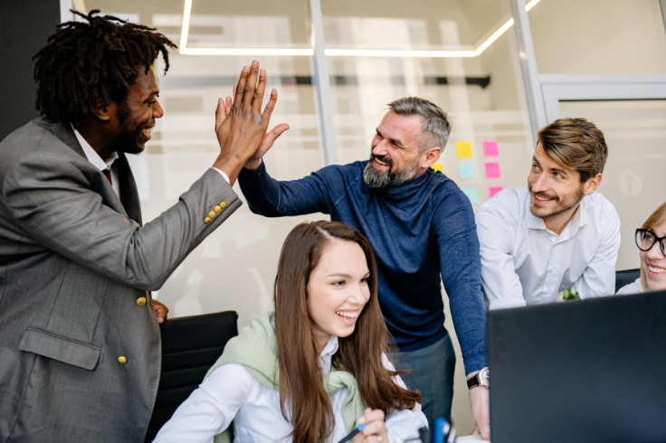 A diverse group of colleagues in an office environment celebrating with a high five and smiles.