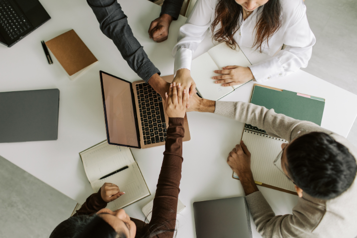 A group of people placing their hands together in a circle over a table with laptops and notebooks.