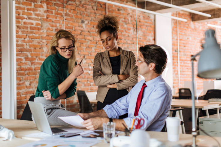 Three marketers are gathered around a desk in an office, looking at documents. 