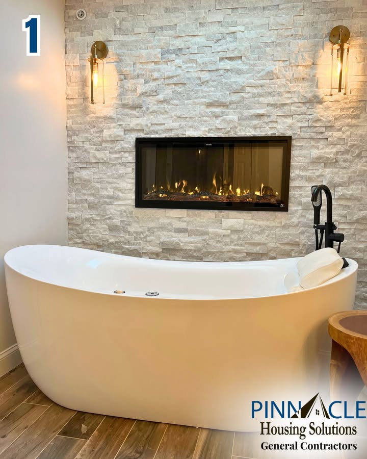 Freestanding white soaking tub in front of a stacked stone wall with a fireplace and two brass sconces in a Chester County, PA bathroom remodel.