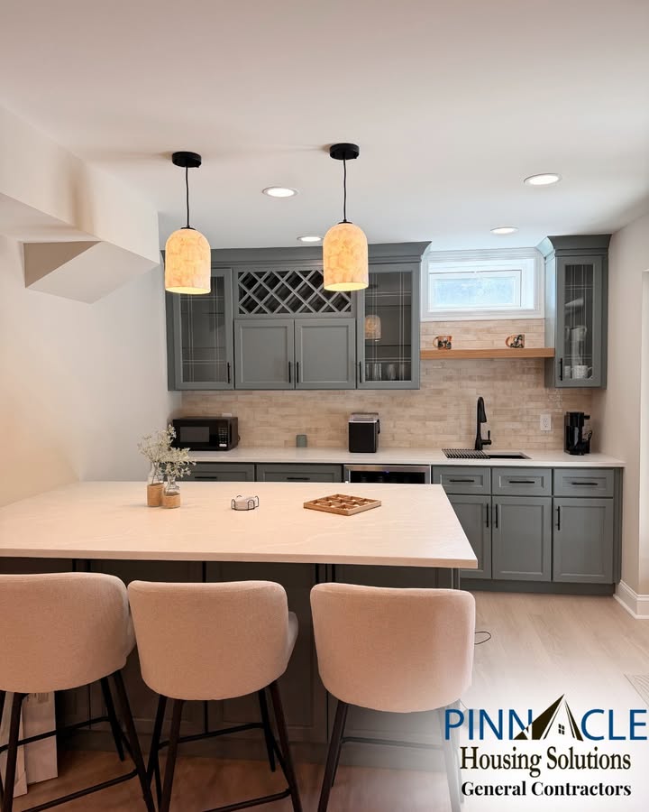 Finished basement bar in Chester County, PA with gray-green glass-front cabinets, a wine rack, two pendant lights, and a peninsula with three barstools.