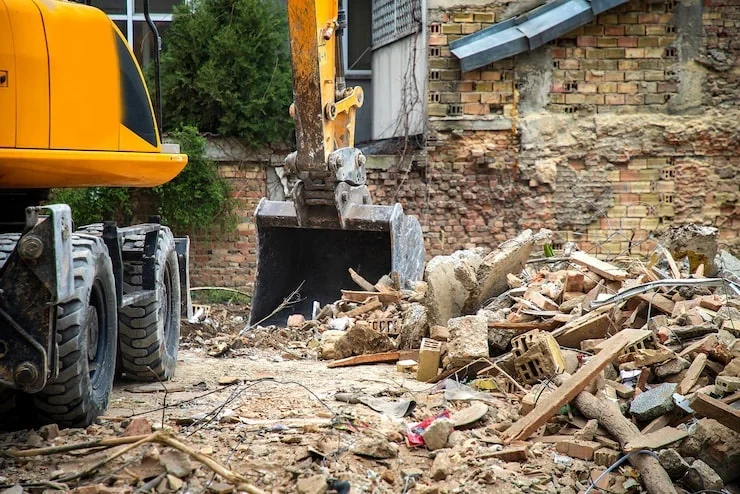 A large yellow excavator bucket and tires next to a pile of rubble from a demolished brick building.