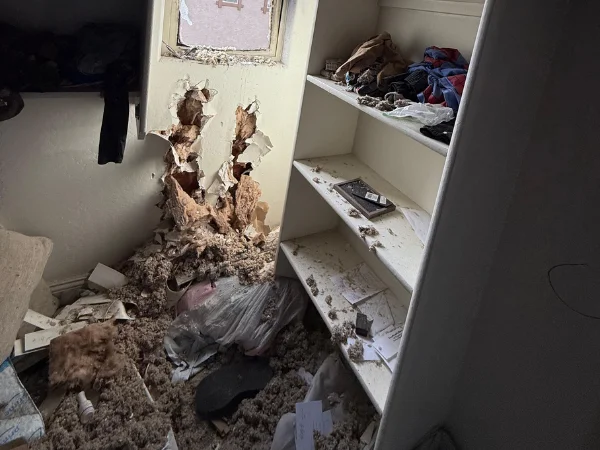 A messy room with a damaged wall exposing insulation, surrounded by debris and items on cluttered shelves.