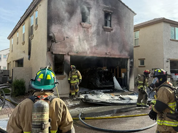 A firefighter in foreground examines a house with a fire-damaged garage and charred vehicle inside, with other firefighters nearby.
