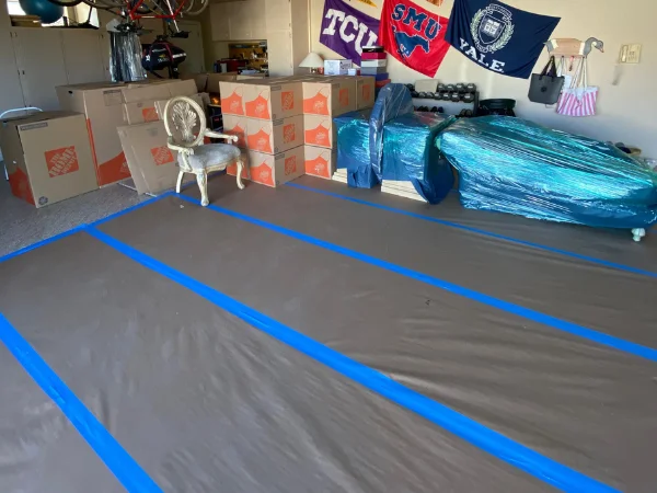 A garage filled with moving boxes, plastic-wrapped furniture, and a drop cloth on the floor with blue tape, ready for moving.
