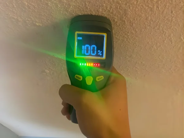A hand holds a digital moisture meter against a textured wall, displaying 100% saturation near a bathroom mirror.