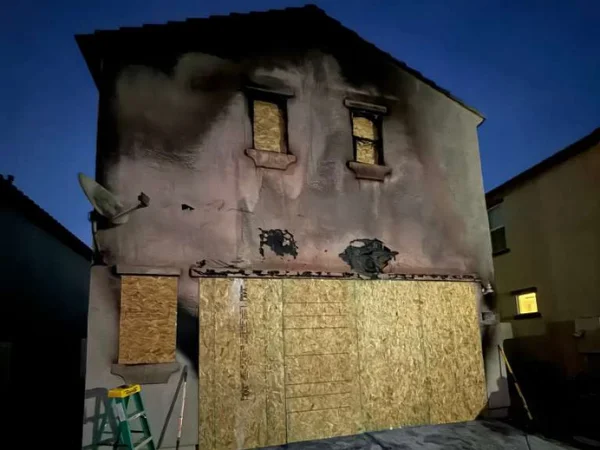 A two-story house with a fire-damaged exterior, boarded-up windows, and black soot marks under a dark sky.