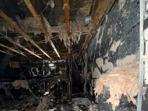 A darkly lit view of a severely fire-damaged building interior with charred walls, exposed wooden beams, and dangling insulation.