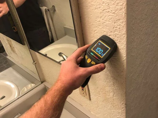 A hand holds a digital moisture meter against a textured wall, displaying 100% saturation near a bathroom mirror.