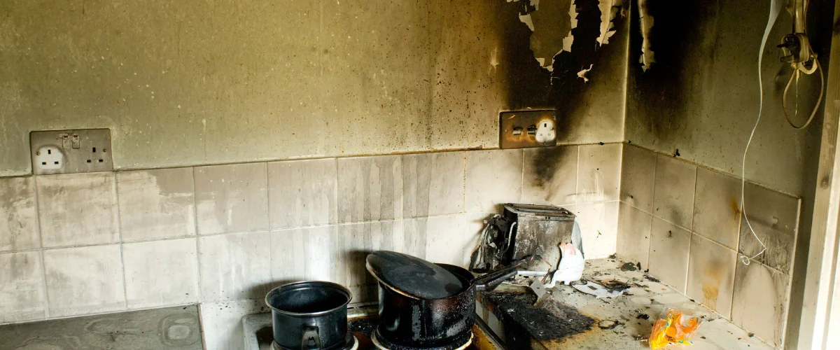 A kitchen extensively damaged by fire, showing blackened walls, charred appliances, and soot-covered surfaces.