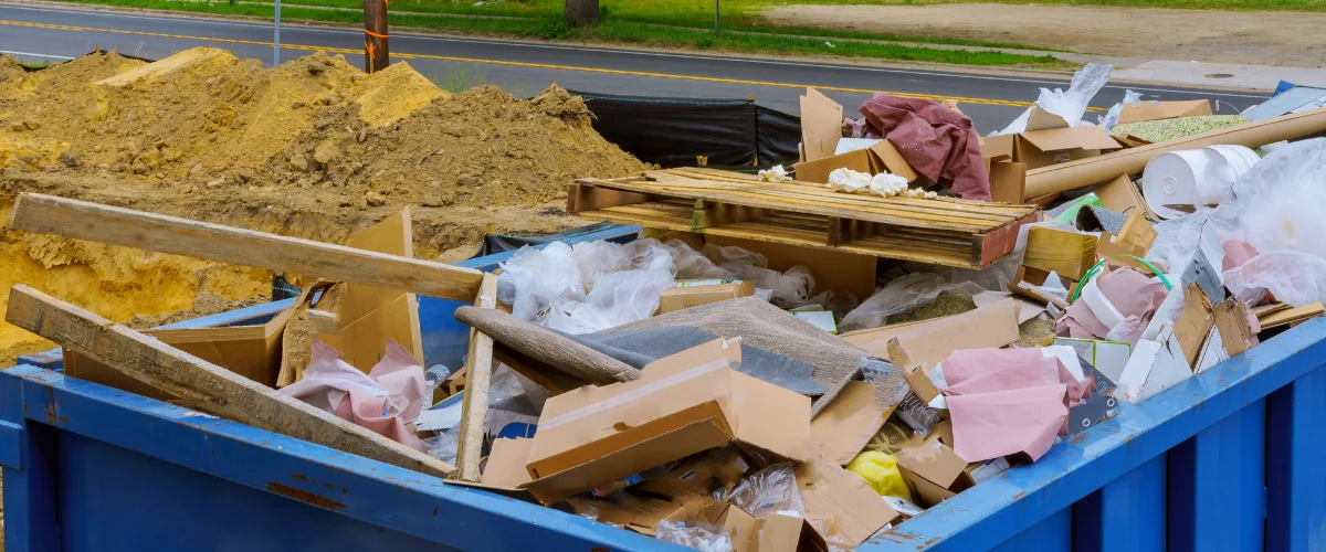 An overflowing blue construction dumpster filled with cardboard, wood, plastic, and debris next to a pile of dirt.