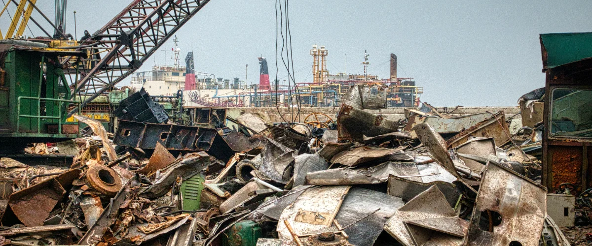 A ship recycling yard with a large pile of rusty metal scrap in the foreground and a large commercial ship in the background.