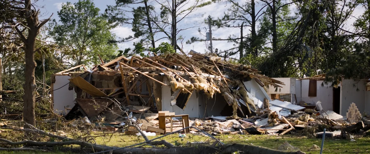 A severely damaged house with its roof completely collapsed, surrounded by debris, broken trees, and insulation from a powerful storm. A severely damaged house with its roof completely collapsed, surrounded by debris, broken trees, and insulation from a powerful storm.