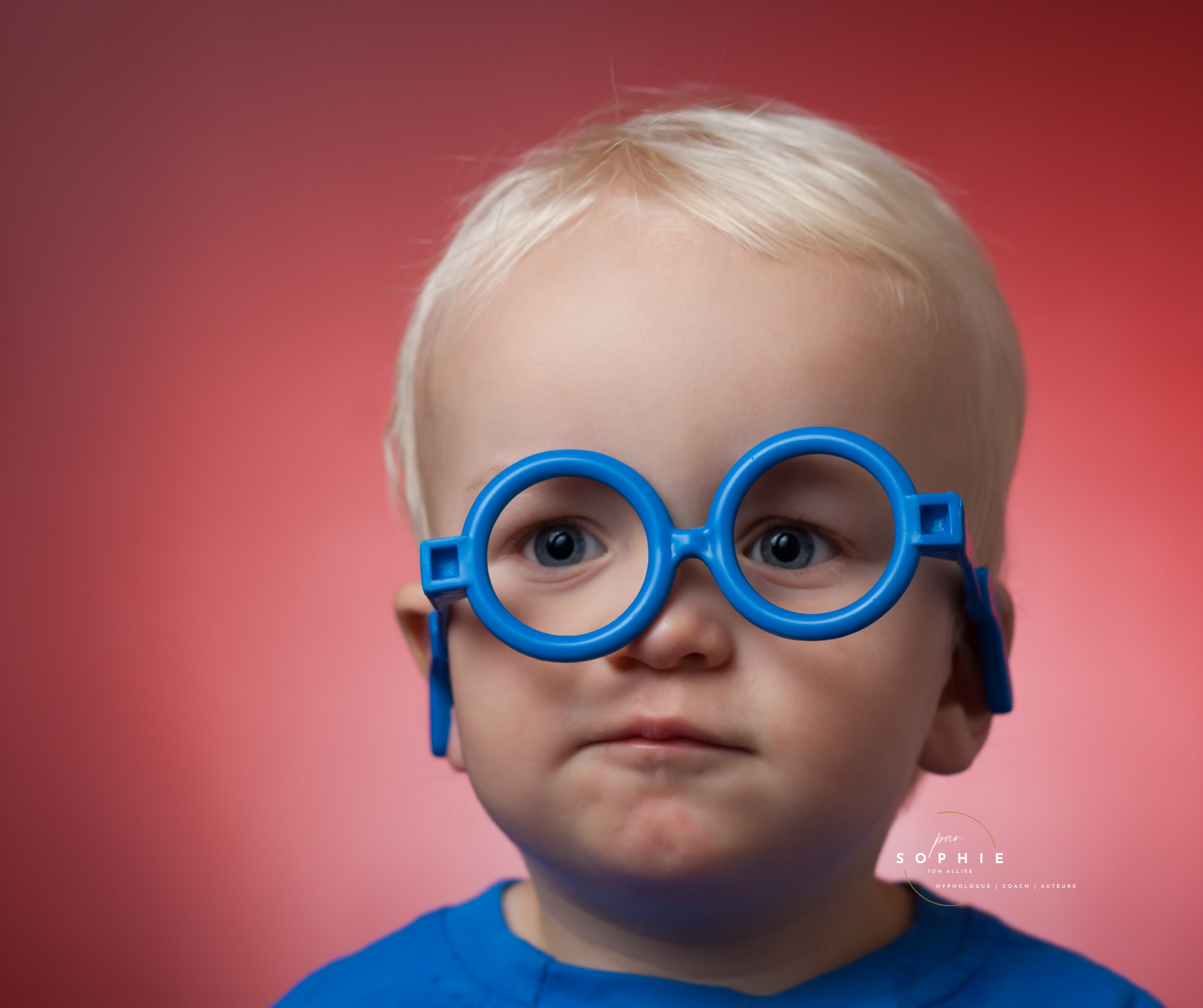 Bébé portant des lunettes jouets bleues, regard curieux, fond rouge Bébé portant des lunettes jouets bleues, regard curieux, fond rouge