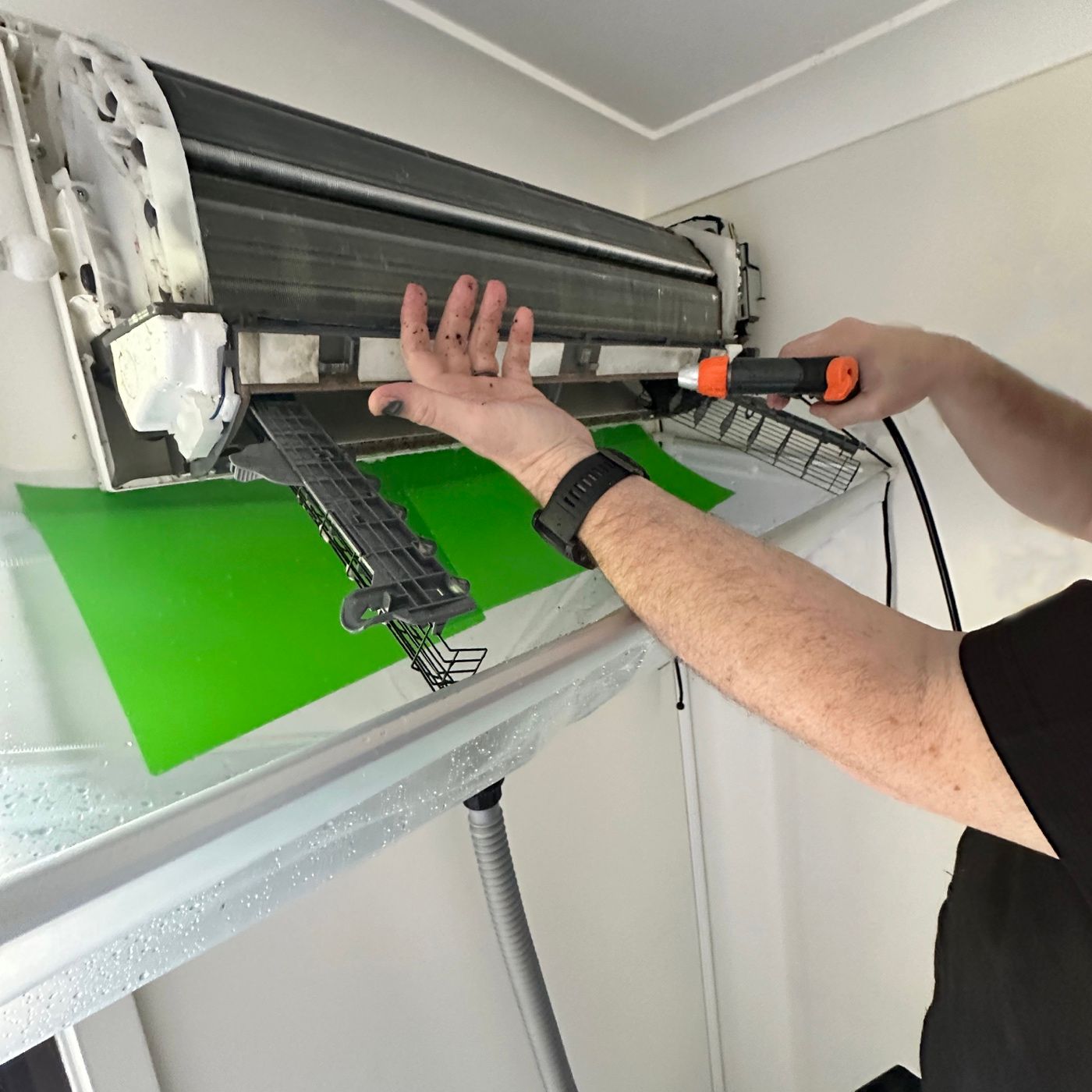 Hands cleaning the coil of an opened split system with a spray gun, green catch bag fitted underneath