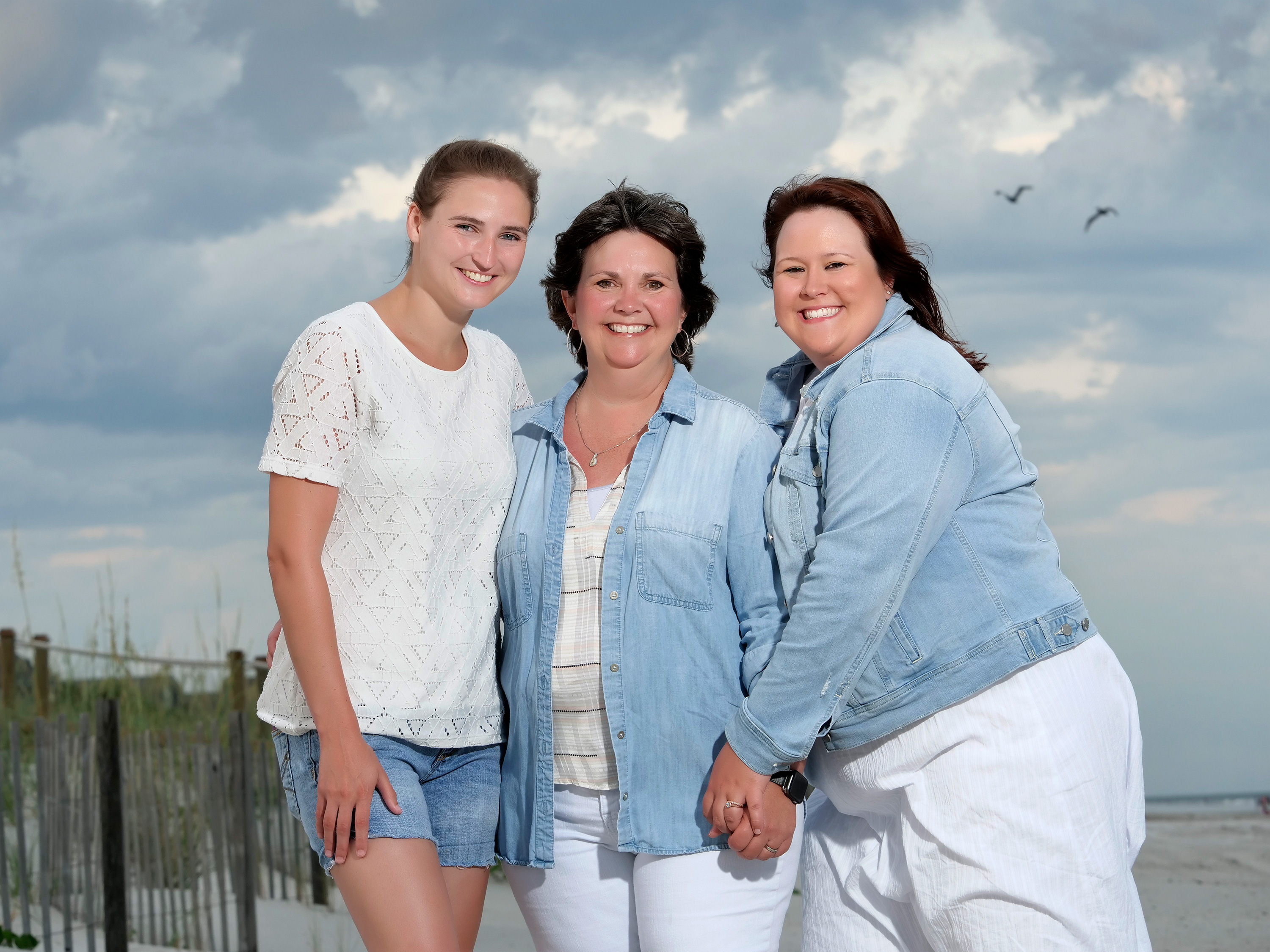 Three women posing together on Hilton Head beach during a family photo session with stormy sky backdrop in Palmetto Dunes
