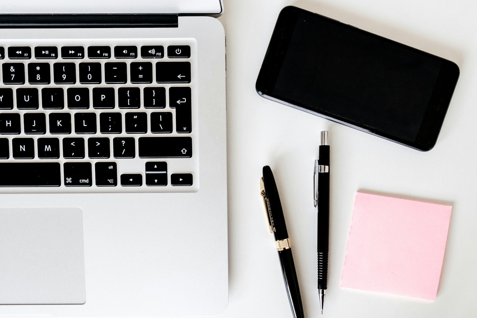A laptop keyboard, a smartphone, two pens and a sticky note on a white desk