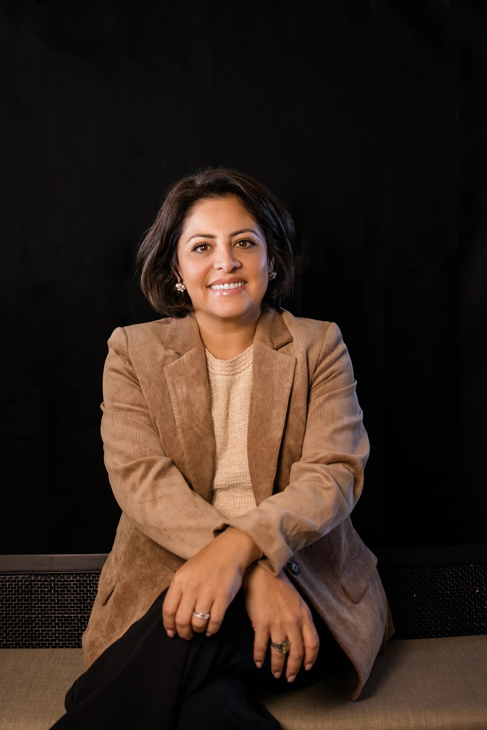 Taryn Pessanha, REALTOR in Calgary, seated portrait in a camel blazer against a dark studio backdrop.