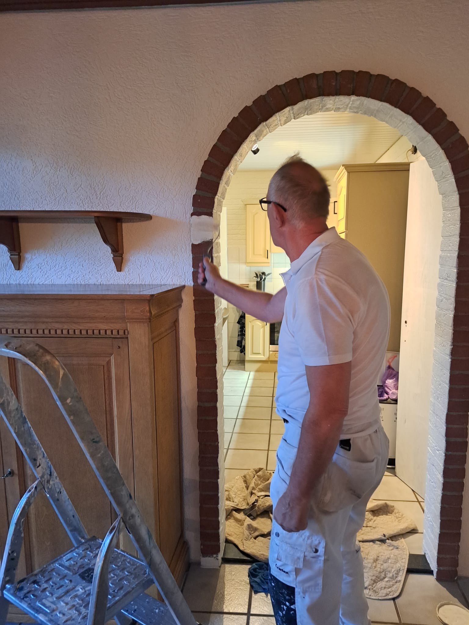 Man schildert een muur in een woning, met een focus op strakke afwerking en details, ladder en schildermaterialen zichtbaar.