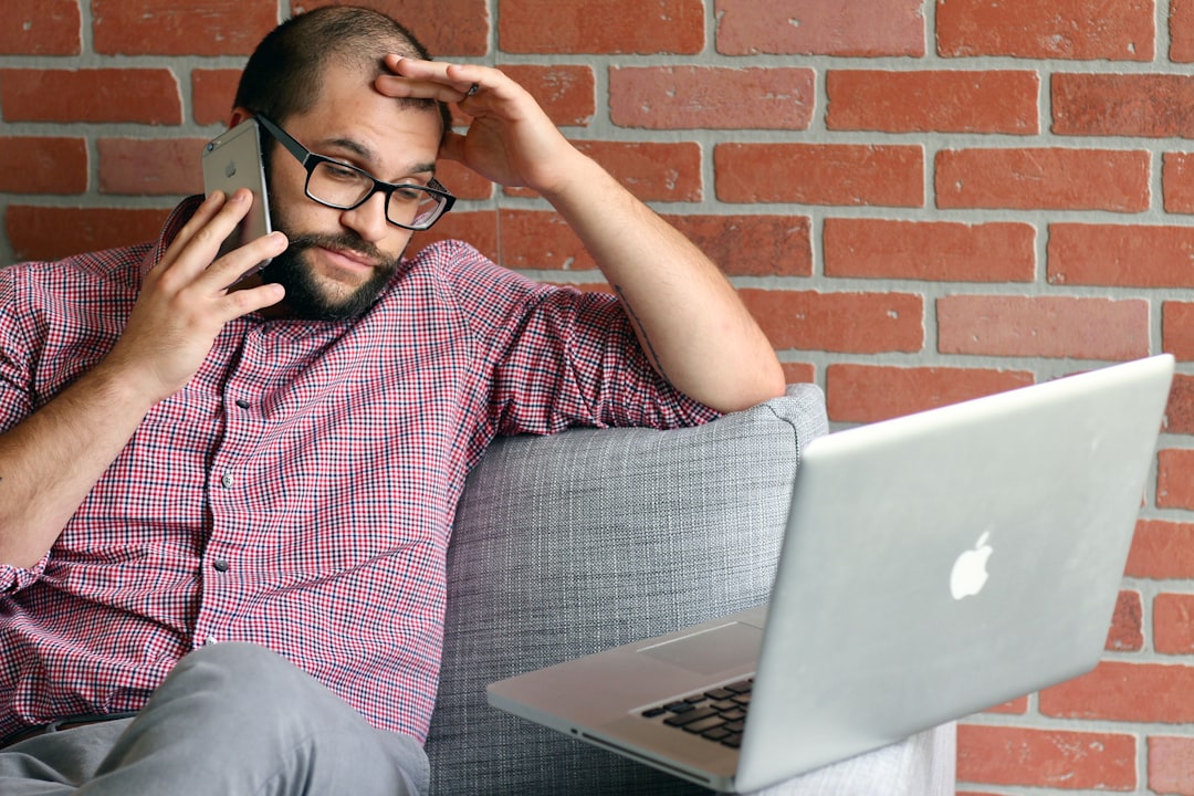 A man looks concerned as he holds a phone to his ear and looks at a lapop.