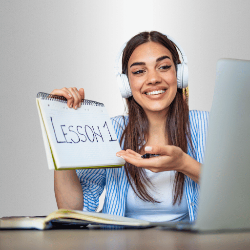 A smiling woman wearing headphones holds up a paper that reads "Lessons" to a laptop camera.