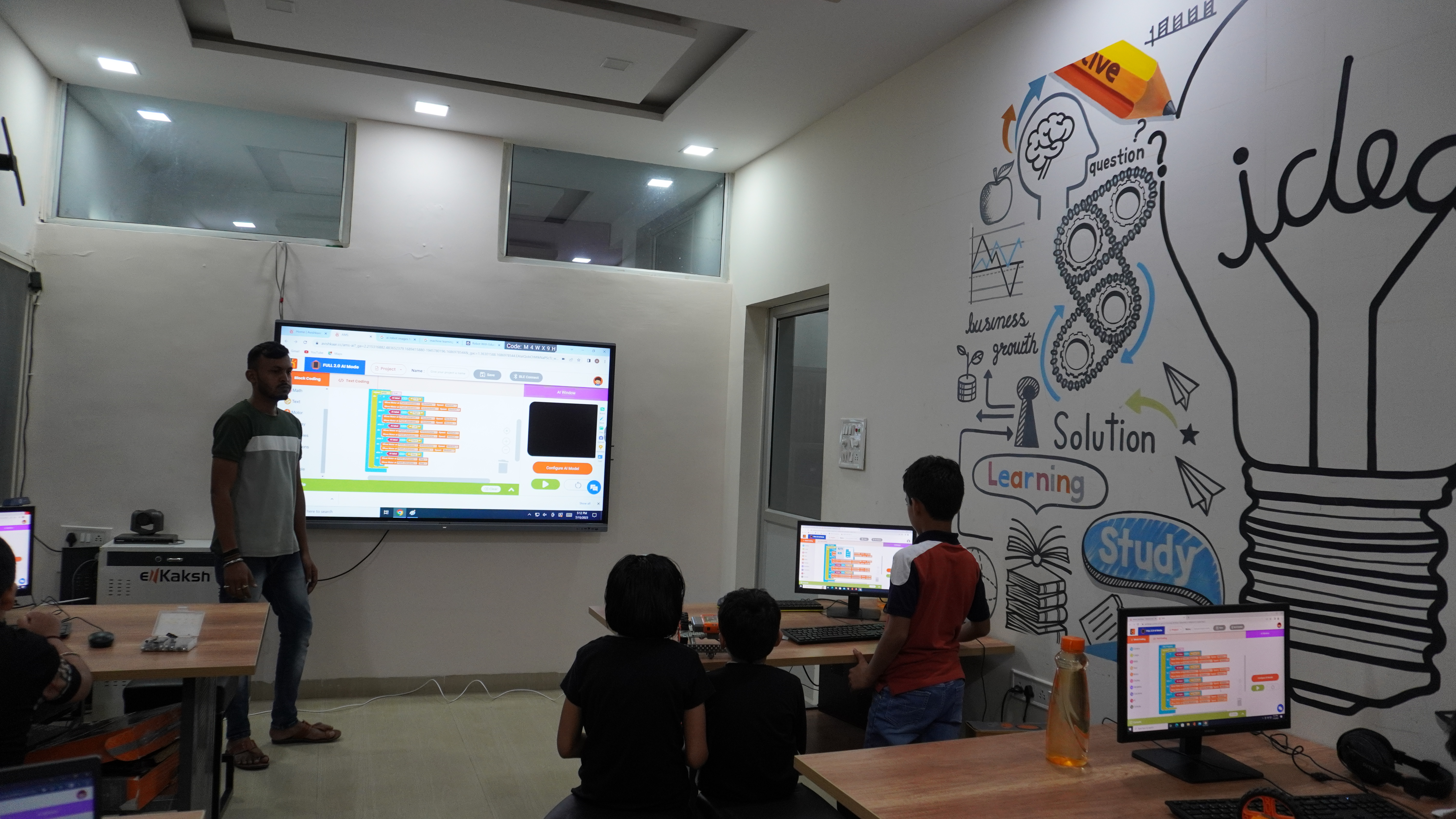 An AVM trainer running AI block-coding on an interactive panel while students code at PCs in a school robotics and AI lab