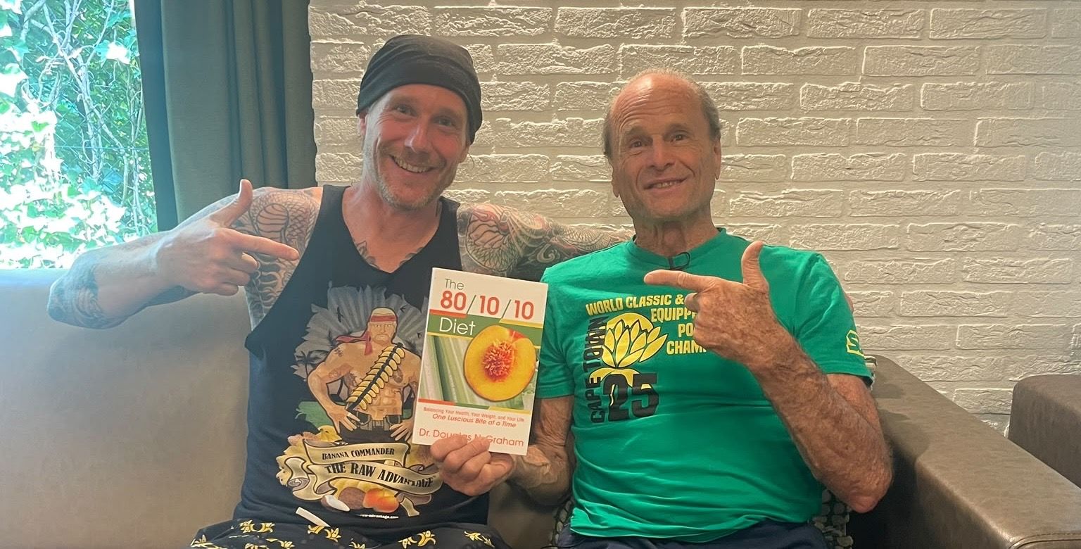 Chris Kendall and Dr. Douglas Graham smiling on a couch, holding a copy of The 80/10/10 Diet