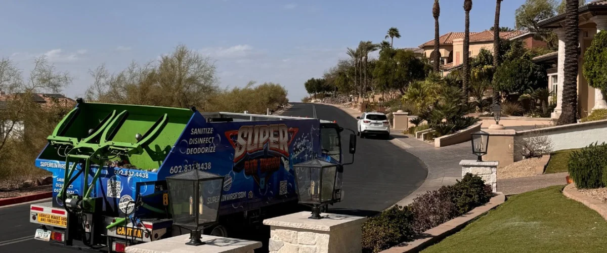 A Super Binwash truck on a residential street in front of upscale homes with palm trees and a white SUV.