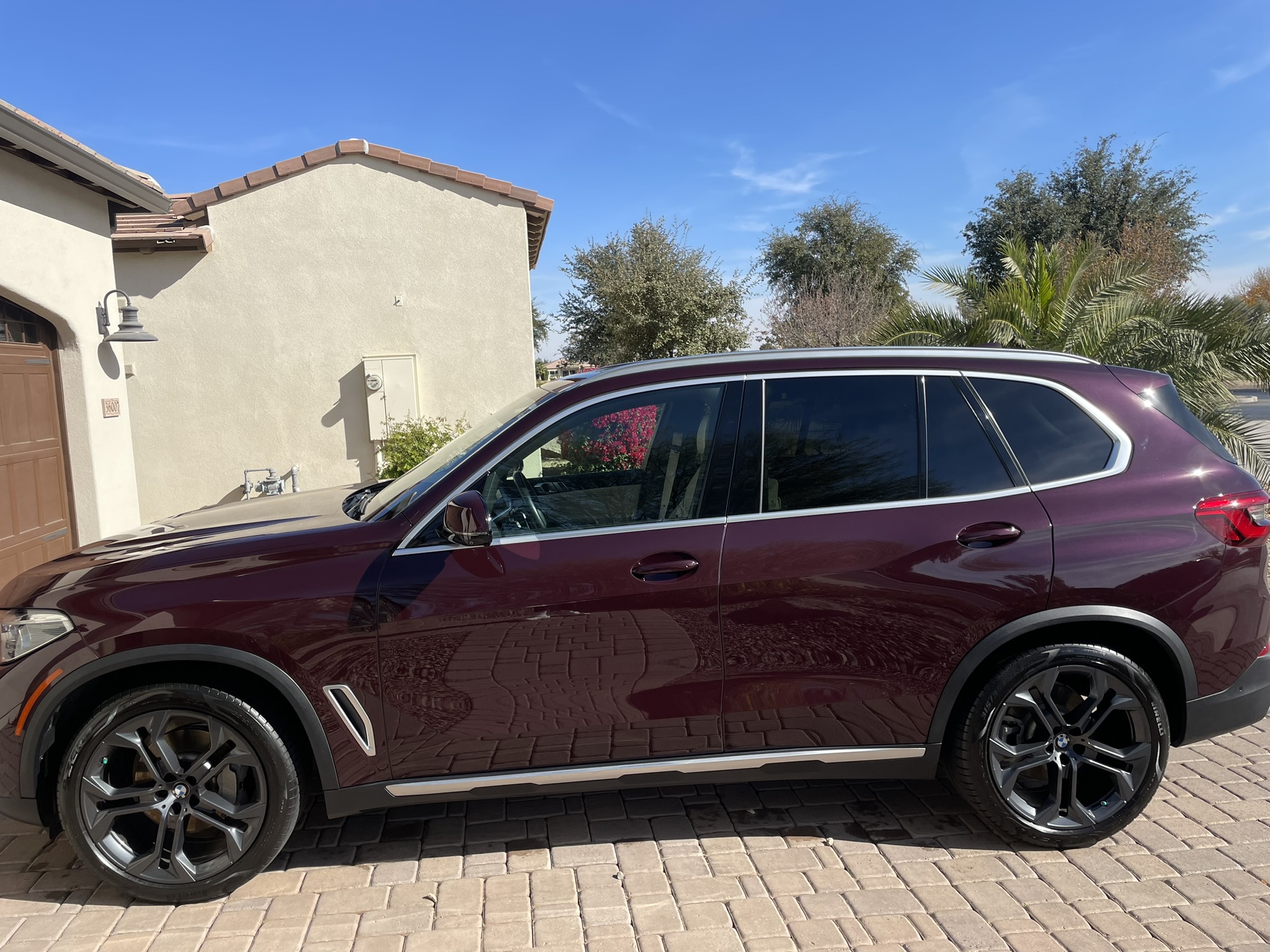 A glossy burgundy BMW X5 SUV parked on a paved driveway, viewed from the side on a sunny day.