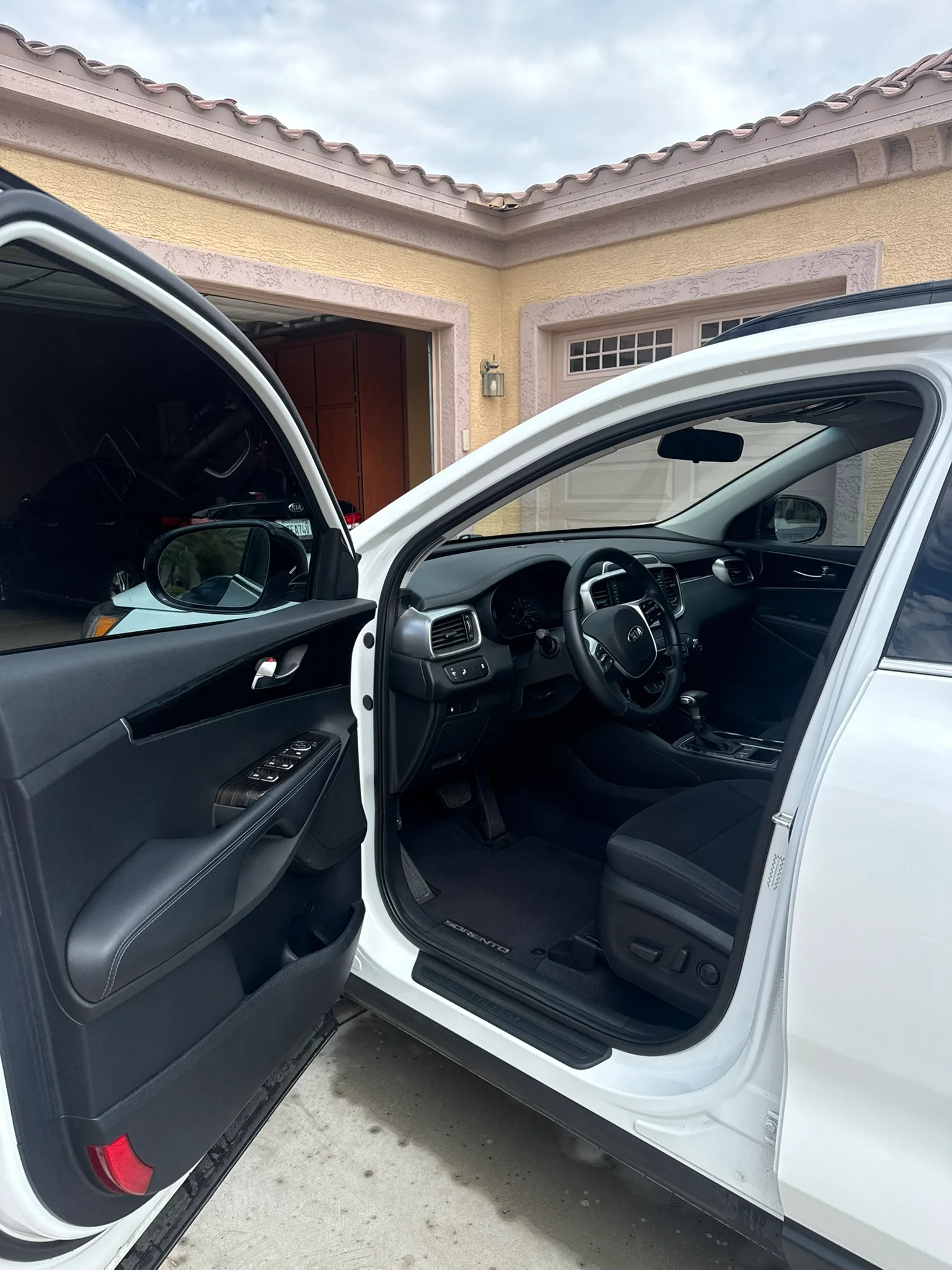 A white Kia Sorento SUV with the driver's door open, revealing the black interior and dashboard, parked outside a garage.