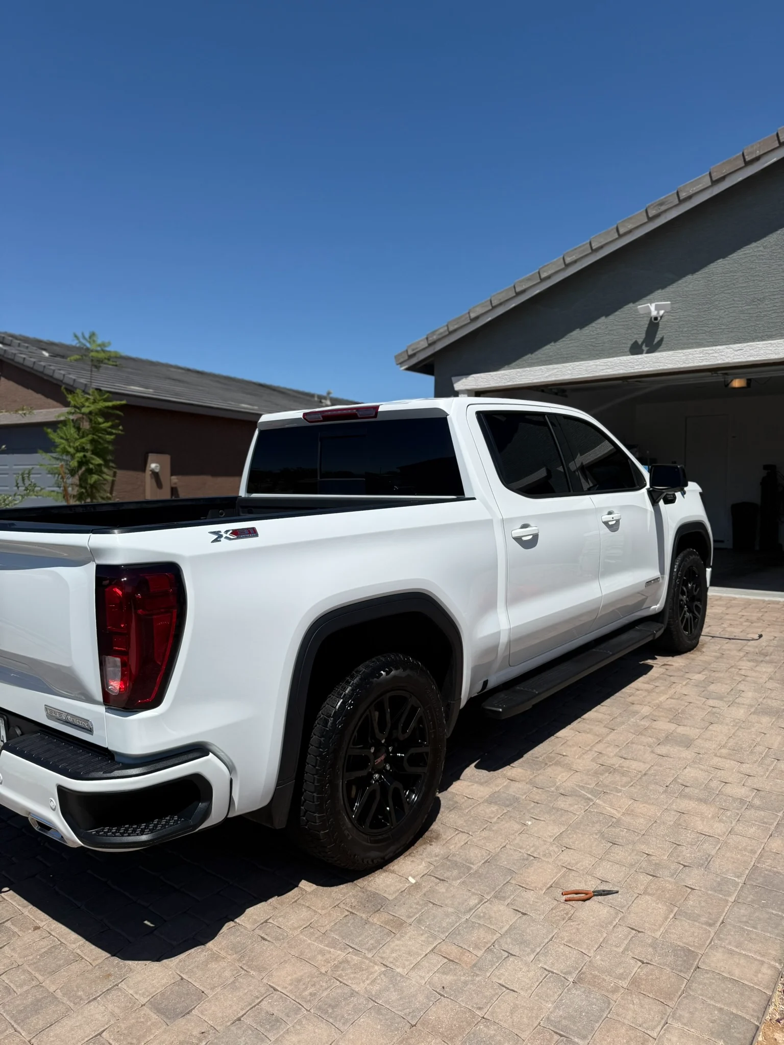 A white GMC Sierra Elevation pickup truck, with black wheels and tinted windows, parked on a paver driveway.