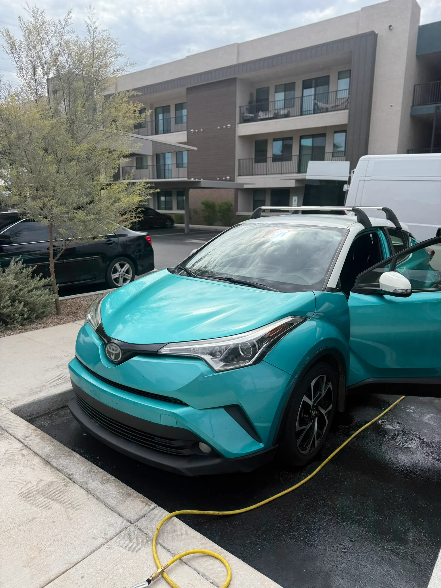 A teal and white Toyota C-HR parked with its driver's door open, a yellow hose on the wet ground nearby.