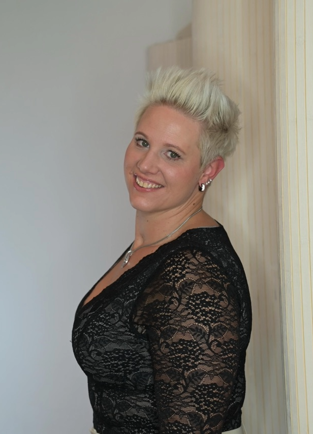 Porträt einer lächelnden Frau mit kurzen blonden Haaren, die seitlich an einer Wand lehnt und ein schwarzes Spitzenoberteil trägt.