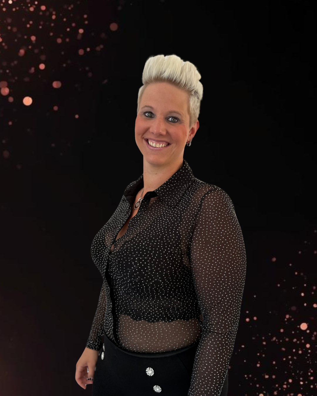Porträt einer lächelnden Frau mit kurzen blonden Haaren, die seitlich an einer Wand lehnt und ein schwarzes Spitzenoberteil trägt.