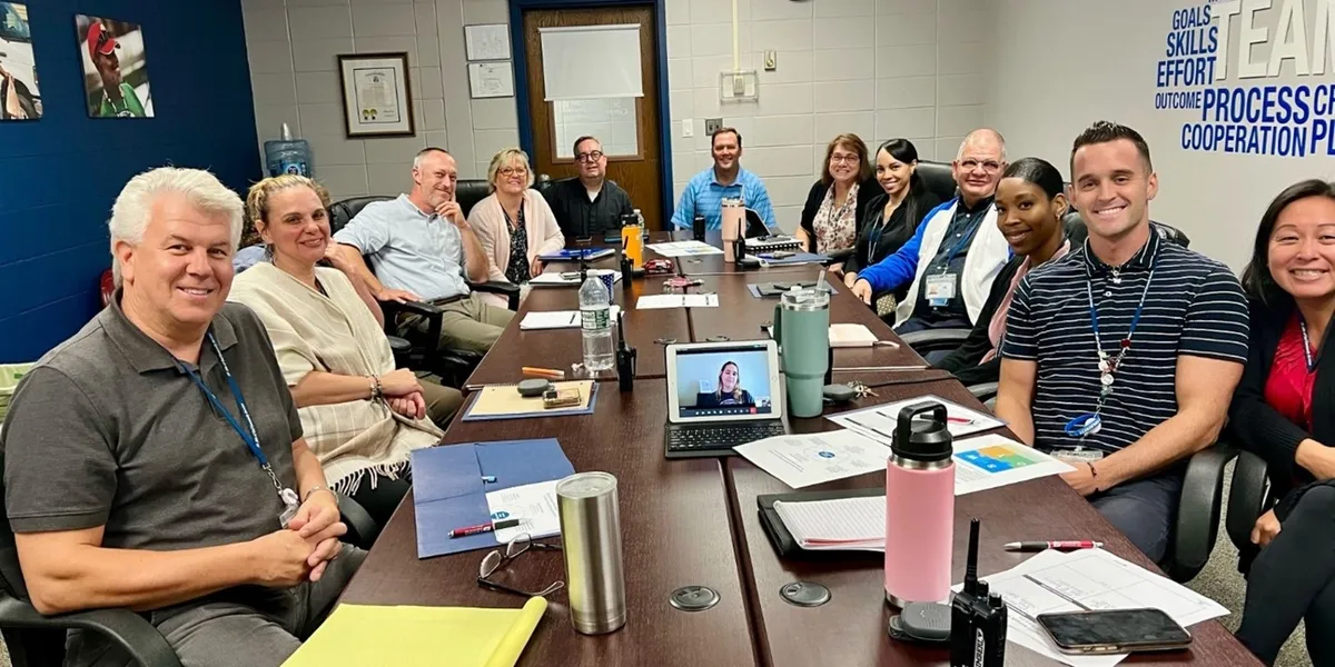 A leadership team gathered around a conference table, with a remote participant joining by video.