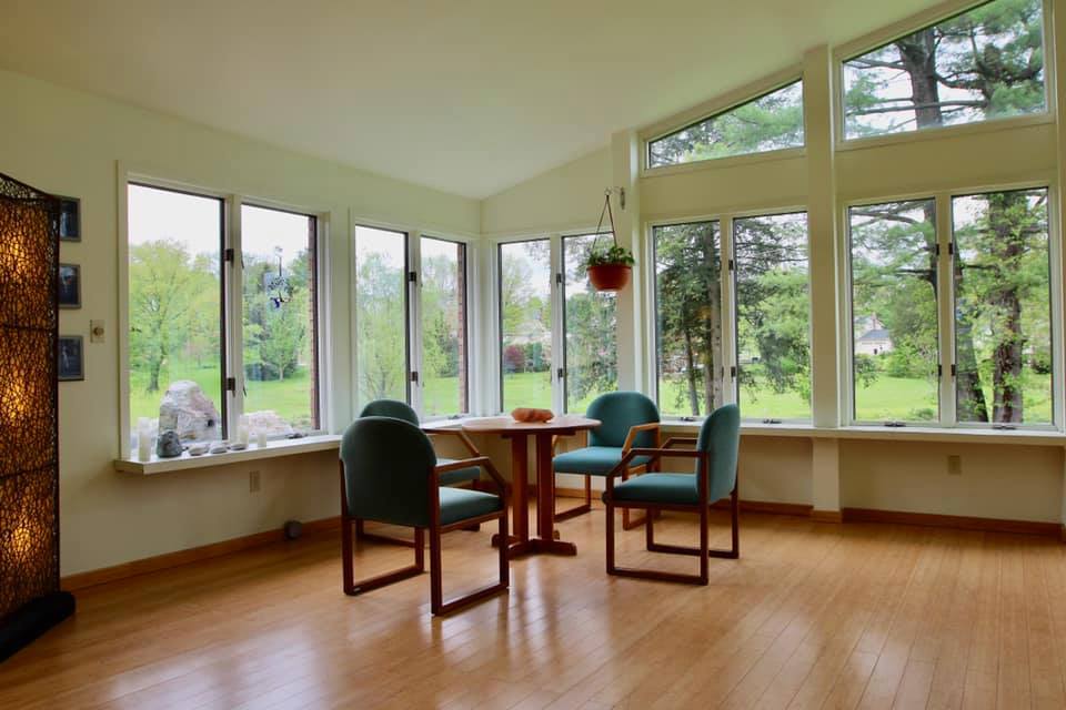 Sunlit meditation nook at Holistic Awakenings with two upholstered chairs, a small table, and wraparound windows opening to trees and a garden
