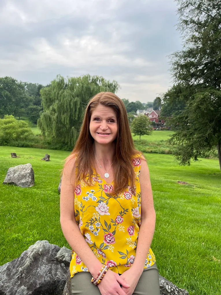 Jenna Galligani, founder of Holistic Awakenings, sitting outdoors in a yellow floral top with willow trees in the background