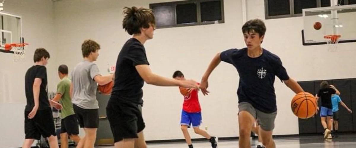 Teen basketball players practicing dribbling and defense drills in a gymnasium.