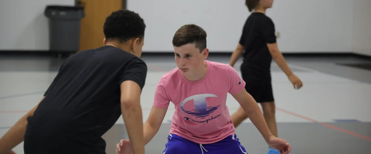 Two young boys in athletic wear face each other, crouched, during a basketball practice or drill inside a gym.