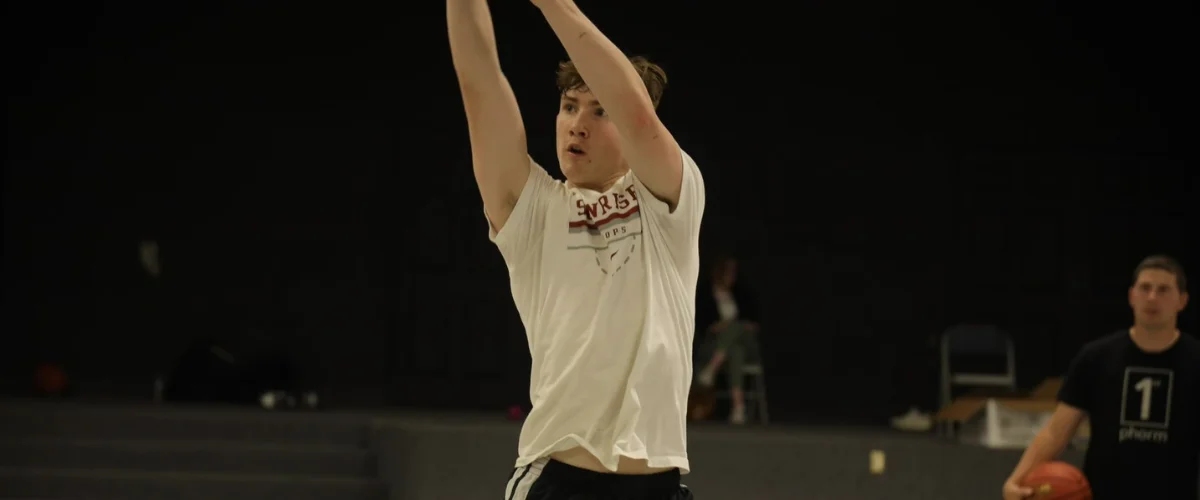 A young male basketball player in a white shirt, arms raised, focused on shooting a ball, with a blurred background. A young male basketball player in a white shirt, arms raised, focused on shooting a ball, with a blurred background.
