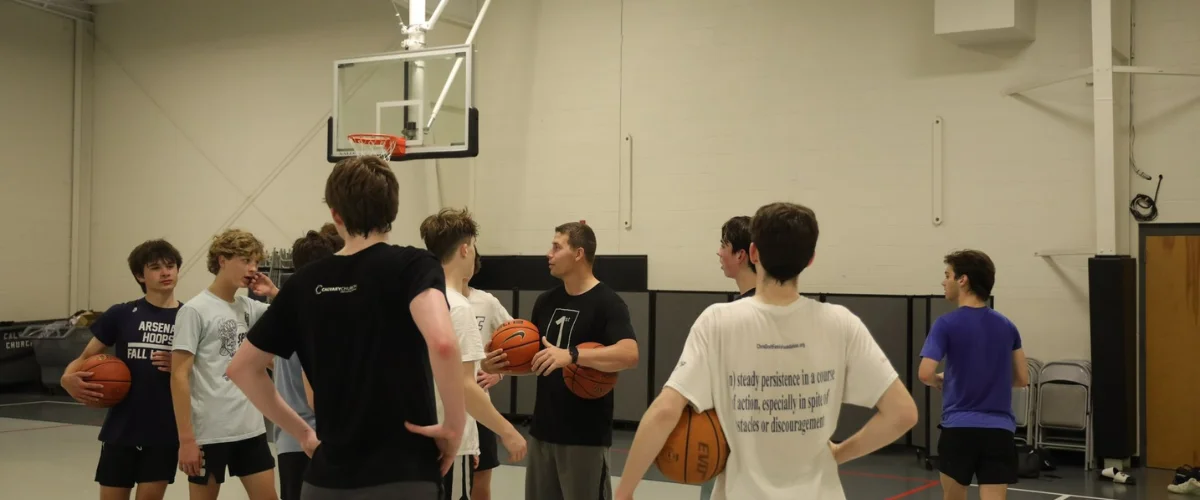 A basketball coach holding two basketballs explains something to a group of teen players in a gym. A basketball coach holding two basketballs explains something to a group of teen players in a gym.