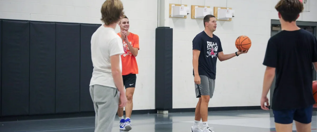 A male basketball coach in a black shirt holds a basketball while instructing young players on the court. A male basketball coach in a black shirt holds a basketball while instructing young players on the court.